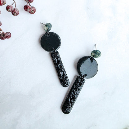 lange zwarte oorbellen met groen oorknopje en zwarte blinkers op marmeren blad met takje