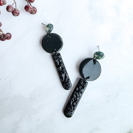 lange zwarte oorbellen met groen oorknopje en zwarte blinkers op marmeren blad met takje