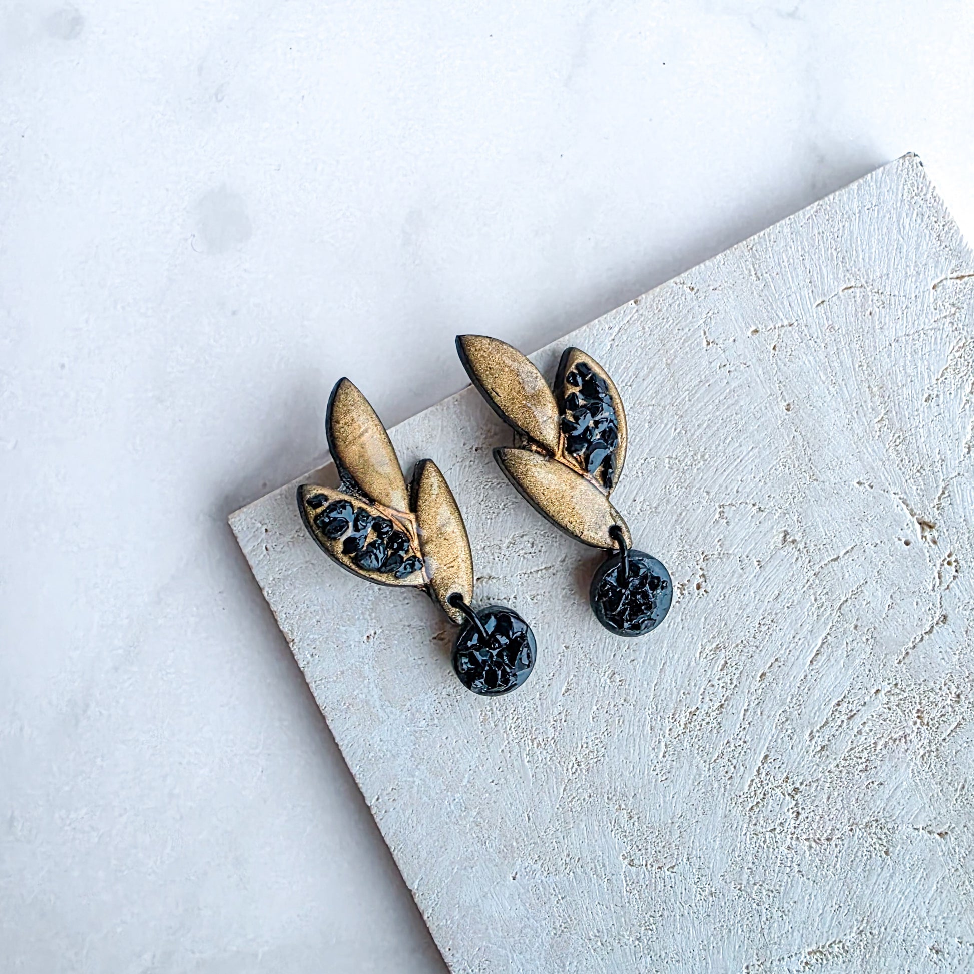 kleine goudkleurige oorbellen in bladvorm met zwarte ronde blinkers op marmeren blad met crème kleurige steen
