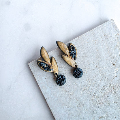 kleine goudkleurige oorbellen in bladvorm met zwarte ronde blinkers op marmeren blad met crème kleurige steen